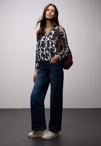 Femme debout portant un chemisier à manches longues à motifs, un jean bleu à jambes larges, des baskets blanches et un sac à bandoulière marron sur un fond uni.