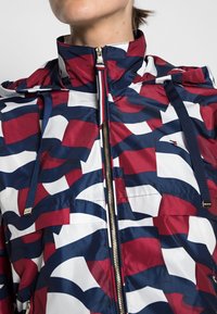Červená, bílá a námořnická vzorovaná bunda s vysokým límcem, zipovým uzavíráním a stahovacími detaily, vyrobená z lehké, lesklé látky.