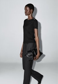 Zwart aansluitend T-shirt met korte mouwen, met knoopaccenten op de schouders, gecombineerd met zwarte tailored broek en een zwarte leren tas.
