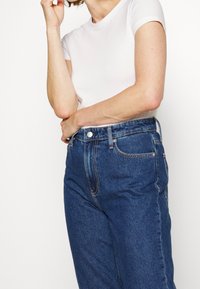 Tummansiniset, korkeavyötäröiset farkut, joissa on napillinen suljenta, etutaskut ja hillitty ompelukuvio, yhdistetty istuvaan valkoiseen t-paitaan.