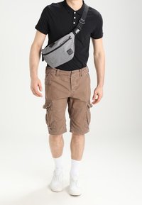Bolso bandolera gris con acentos negros, llevado sobre una camiseta polo negra. Shorts cargo marrones con bolsillos laterales. Zapatillas blancas y calcetines hasta el tobillo.