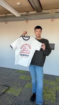 Jonge man staat binnen en houdt een wit T-shirt vast met rode en zwarte tekst waarop staat "Indigo Uniforms Replay Denim Goods."