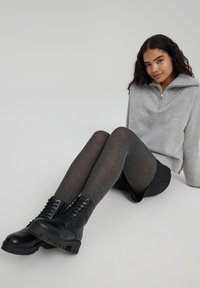 Stivali neri con lacci e suole spesse, collant grigi a coste e un maglione grigio chiaro con zip, indossati su uno sfondo neutro.