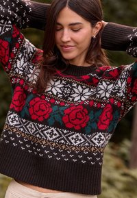 Pull en maille avec une base marron foncé, des roses rouges et des flocons de neige blancs. Poignets et ourlet côtelés, avec des motifs géométriques partout.