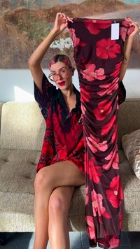 Avondjurk met bloemenprint op een donker bordeauxrode ondergrond, met grote roze en rode bloemen en zijslingers. Model houdt jurk omhoog ter display.