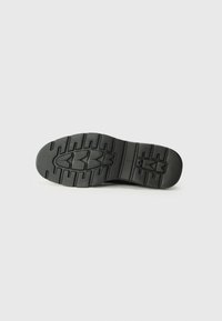 Semelle de chaussure noire avec des motifs de crampons profonds, présentant des rainures angulaires et une conception robuste, fabriquée en caoutchouc pour une meilleure adhérence et durabilité.