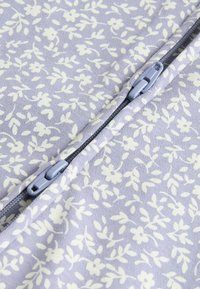 Tissu violet clair avec un motif floral crème. Présente une fermeture éclair centrale avec un matériel gris, améliorant la fonctionnalité et le design.