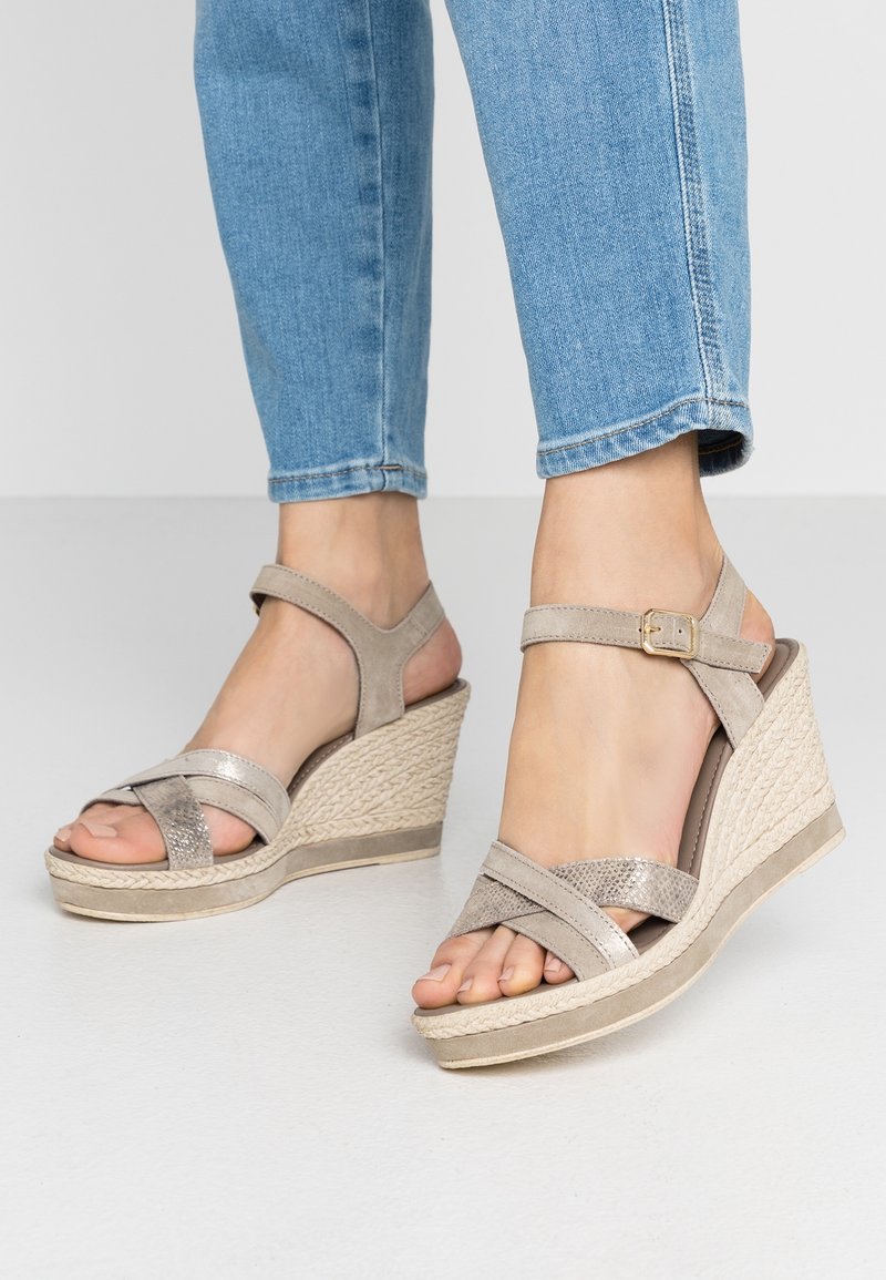 Sandales compensées beiges avec un design de brides croisées texturées et une boucle à la cheville, présentant une semelle intermédiaire tissée et une finition lisse.