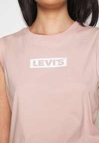 Růžové bezrukávové tričko vyrobené z bavlny, s bílým logem Levi's na obdélníkovém pozadí. Hladká textura, kulatý výstřih.