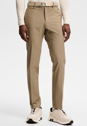 Homme portant un pantalon beige slim, une ceinture tressée claire, une chemise blanche à manches longues et des baskets crème avec des touches noires.