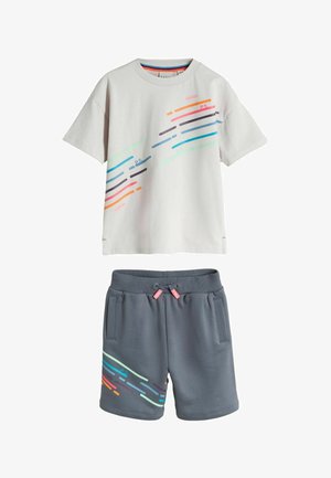 Weißes T-Shirt mit kurzen Ärmeln und bunten diagonalen Streifen, kombiniert mit grauen Shorts mit Kordelzug, die am Bein passende Streifenmuster aufweisen.