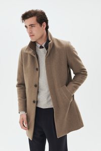 Manteau en laine marron avec un col montant, une fermeture boutonnée sur le devant et des poches latérales, porté sur un pull gris clair et un pantalon foncé.