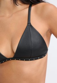 Top bikini triangolare in pelle nera con spalline sottili, caratterizzato da borchie dorate lungo il sottofasciato. Texture liscia e design minimalista.