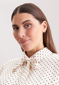 Crème blouse met zwarte stippen, voorzien van een strik aan de kraag. Glad textuur, gestructureerde halslijn en korte mouwen zichtbaar.
