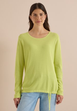 Kvinde med langt mørkt hår iført en lysegrøn langærmet sweater og lyseblå jeans, stående mod en ensfarvet beige baggrund.