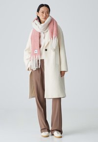 Cappotto di lana crema con bottoni neri, maglione beige, sciarpa rosa e bianca con frange, pantaloni marroni su misura e sneakers color camel.