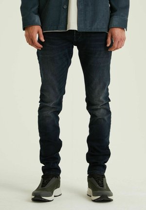 Dunkelblaue Slim-Fit-Jeans mit einem verblassten Design. Kombiniert mit grauen Sneakers, die eine auffällige weiße Sohle und schwarze Schnürdetails aufweisen.