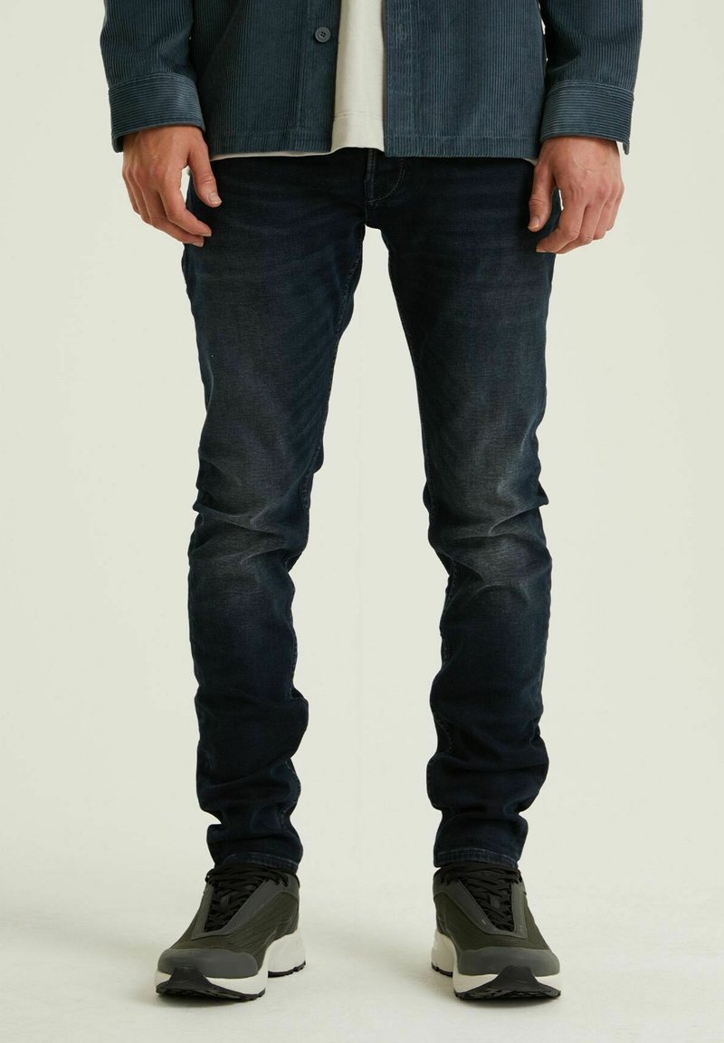 Donkerblauwe slim-fit jeans met een vervaagde textuur. Gepaard met grijze sneakers met een opvallende witte zool en zwarte veters.