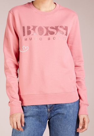 Osoba ubrana w różową bluzę z długim rękawem z napisem "BOSS HUGO BOSS" i niebieskie dżinsy na jednolitym tle.