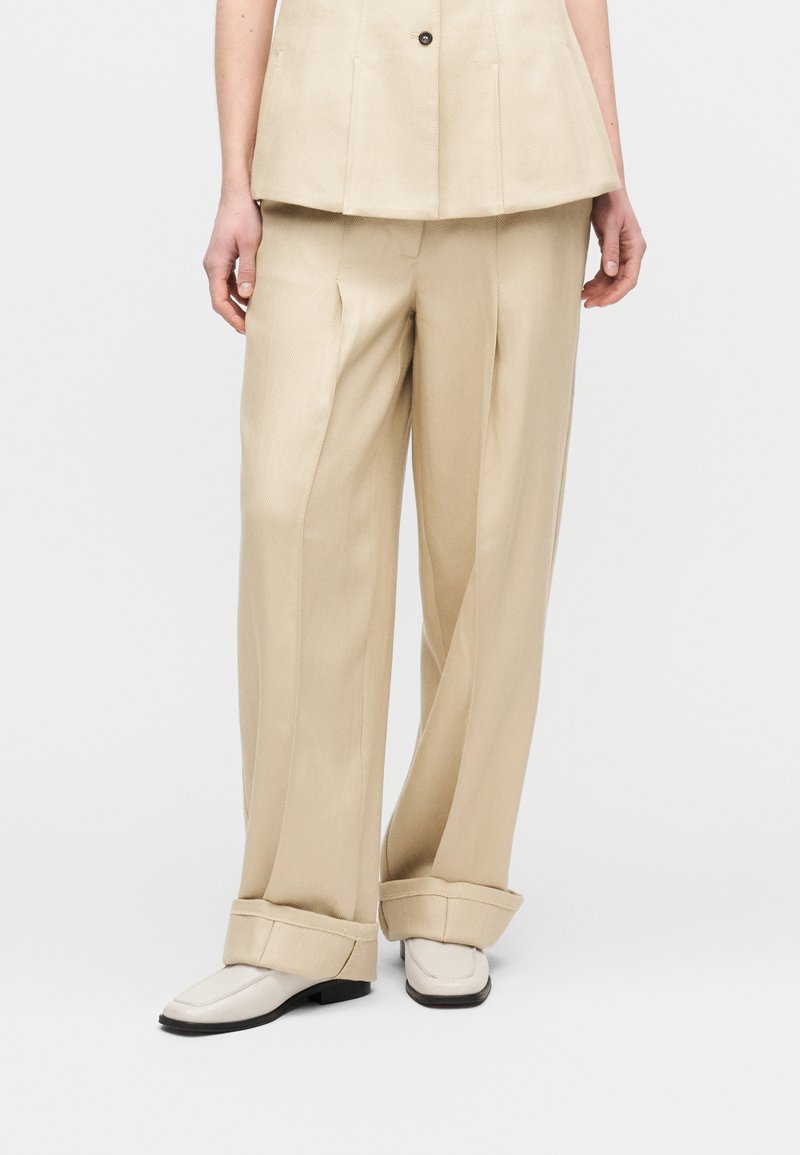 Άτομο που φοράει μπεζ παντελόνι φαρδιά με μανσέτες, αντίστοιχο πουκάμισο με κουμπιά και λευκά μοκασίνια σε απλό λευκό φόντο.