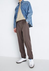 Denimjack, beige top, bruine broek en witte sneakers met blauwe accenten. Opvallende kenmerken: ontspannen pasvorm, knoopdetails en contrasterende texturen.
