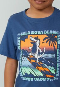 Niebieska bawełniana koszulka z kolorową grafiką krokodyla surfującego, falami i palmami, z napisem "Cala Nova Beach".