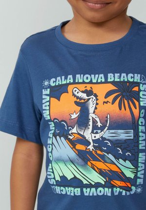 Niebieska bawełniana koszulka z kolorową grafiką krokodyla surfującego, falami i palmami, z napisem "Cala Nova Beach".