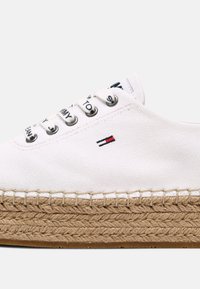 Espadrille blanche en toile avec semelle épaisse en jute tressée, œillets argentés, lacets blancs de marque et petit logo rouge et marine sur le côté.