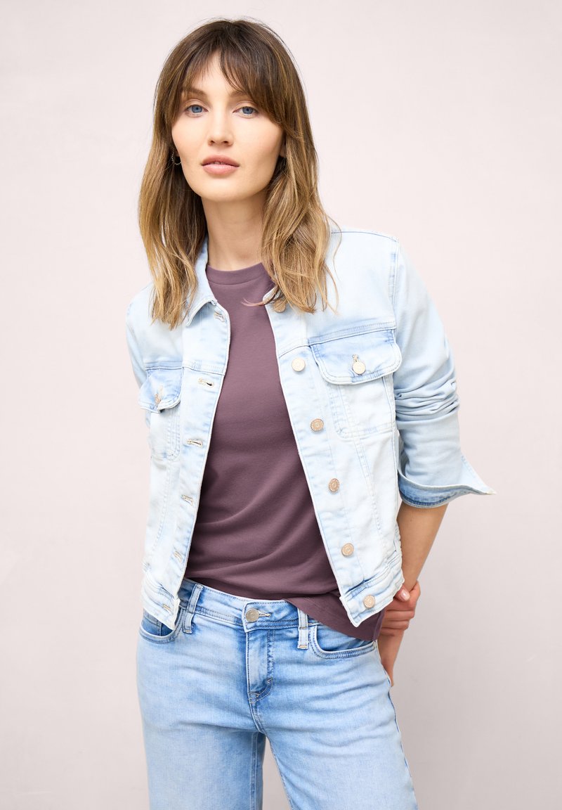 Jeune femme portant une veste en denim bleu clair par-dessus un t-shirt violet et un jean bleu clair, debout devant un fond uni.