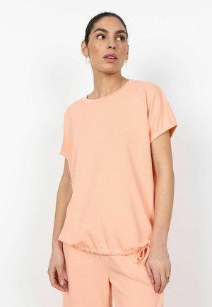 Donna che indossa una maglietta pesca a maniche corte con orlo con coulisse e pantaloni abbinati, in piedi contro uno sfondo bianco, che guarda di lato.