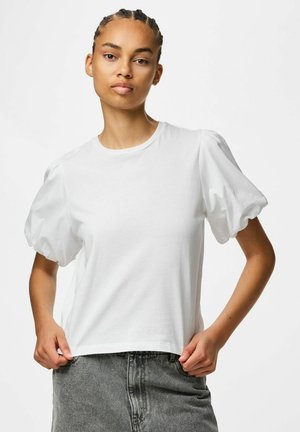 Jeune femme aux cheveux tressés portant un haut blanc à manches bouffantes et un jean gris sur fond blanc uni.