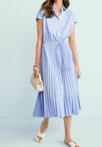 Niebieska i biała sukienka koszulowa w paski z zapięciem na guziki, krótkimi rękawami, ściągniętą w talii i plisowanym dołem. W komplecie z beżową torebką.