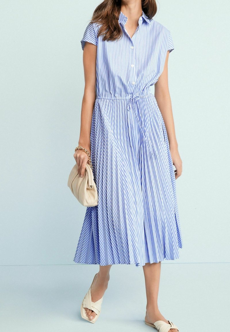 Niebieska i biała sukienka koszulowa w paski z zapięciem na guziki, krótkimi rękawami, ściągniętą w talii i plisowanym dołem. W komplecie z beżową torebką.