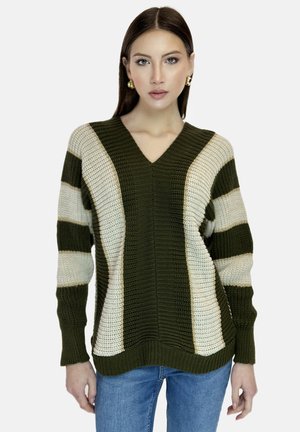 Kvinde iført en grøn og beige stribet strikket trøje med V-hals og blå jeans, stående mod en ensfarvet hvid baggrund.