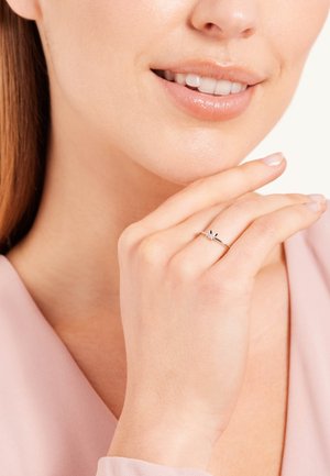 Silver ring with delicate design featuring a central gemstone. Smooth band, worn on a hand with light nail polish, near a softly lit face.