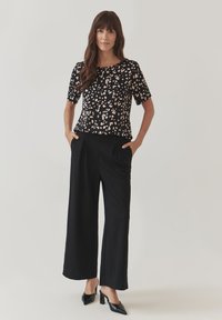 Top nero con un motivo a pois rosa e bianco, abbinato a pantaloni neri larghi e scarpe col tacco nere. Outfit casual.