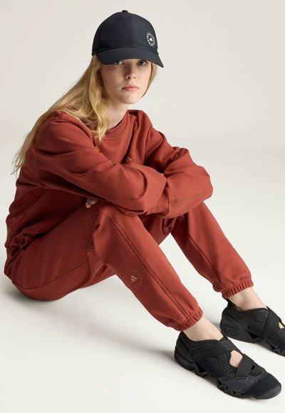 Młoda kobieta siedząca na podłodze, ubrana w rdzawo-brązową bluzę i spodnie dresowe, czarną czapkę oraz czarne siateczkowe trampki na rzepy.