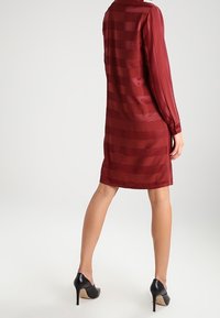 Bordowa satynowa sukienka z długimi rękawami, z poziomymi paskami. Noszona z czarnymi szpilkami. Prosty, do kolan fason.