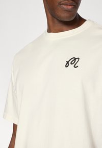 Μπλούζα από βαμβάκι σε κρεμ χρώμα με στρογγυλή λαιμόκοψη, που φέρει ένα μαύρο κεντημένο λογότυπο "m" στο αριστερό στήθος. Κοντά μανίκια, λεία υφή.