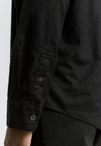 Zwarte langekous-shirt met knoopmanchetten, een effen textuur en subtiele stikdetails langs de zoom en de mouwrand.