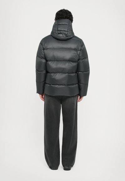 Schwarze Puffjacke mit Kapuze, die ausgeprägte horizontale Steppnähte und eine lockere Passform aufweist. Getragen über weit geschnittenen grauen Hosen.