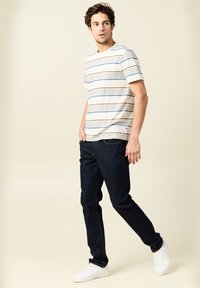 Raidallinen lyhythihainen t-paita, jossa on sinisiä ja ruskeita vaakasuoria viivoja, yhdistettynä tummiin farkkuihin ja valkoisiin tennareihin, joita käyttää miesmalli.