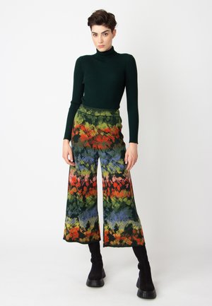 Groene geribbelde coltrui gecombineerd met een wijdlopende broek met een meerkleurig bloemenpatroon in tinten van rood, groen, blauw en zwart.