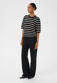 Černobíle pruhovaný svetr s krátkými rukávy, vyrobený z měkkého materiálu. V kombinaci s černými širokými kalhotami a černými botami s špičatou špičkou.