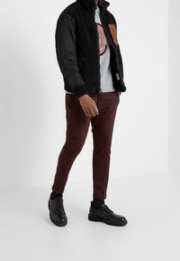 Chaqueta bomber de forro polar y nylon negra con un parche marrón, sudadera gris, pantalones ajustados burdeos y zapatos negros con cordones.