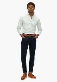 Lichtgroen overhemd met knopen en een zak, donkerblauwe jeans, een brown leren riem en bruine schoenen. Model staat met de handen op de heupen tegen een witte achtergrond.