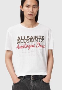 Άνδρας με μακριά σκούρα μαλλιά που φοράει λευκή μπλούζα με επαναλαμβανόμενο κείμενο "ALLSAINTS" και κόκκινη γραφική επιγραφή "Analogue Days", μαύρη ζώνη και σκούρο τζιν.