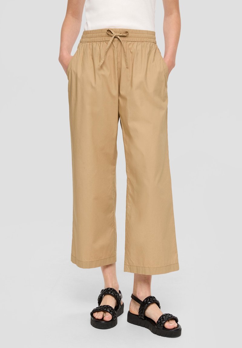 Personne portant un pantalon large beige court avec une taille à cordon de serrage et des sandales compensées noires tressées.
