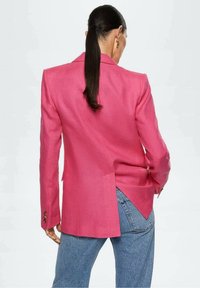 Femme avec une longue queue de cheval sombre portant un blazer rose vif avec une fente à l'arrière, associée à un jean bleu, debout devant un fond blanc.