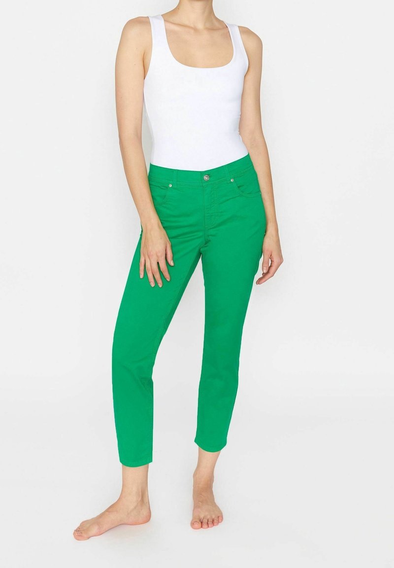 Femme portant un haut blanc sans manches et un pantalon slim vert vif longueur cheville, debout pieds nus sur fond blanc.